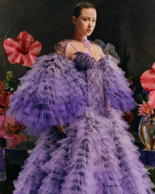Woman in ruffled purple couture gown poses in studio with floral props and dark backdrop for fashion editorial portrait.