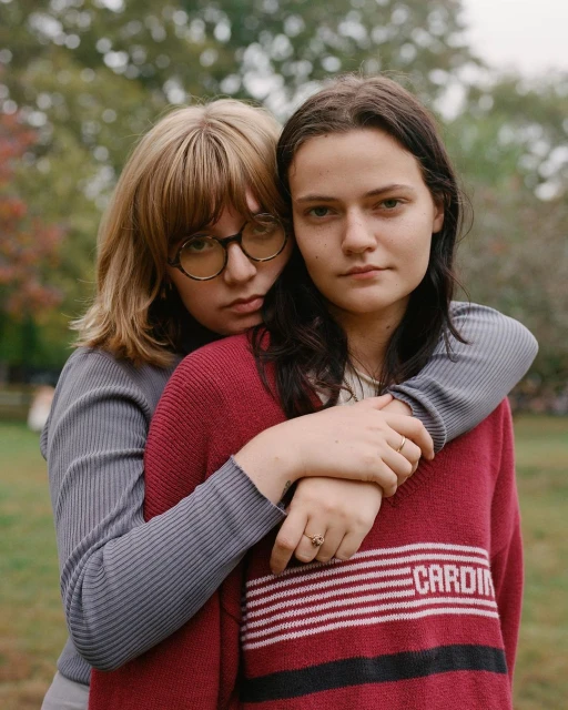 Portrait of two adult females embracing outdoors. One with blonde hair in glasses and one with dark hair, both wearing sweaters.