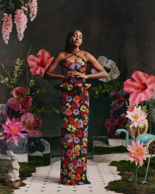 Woman in floral dress poses amid lush flower garden setting, captured in a studio with artificial lighting, creating an editorial fashion image.