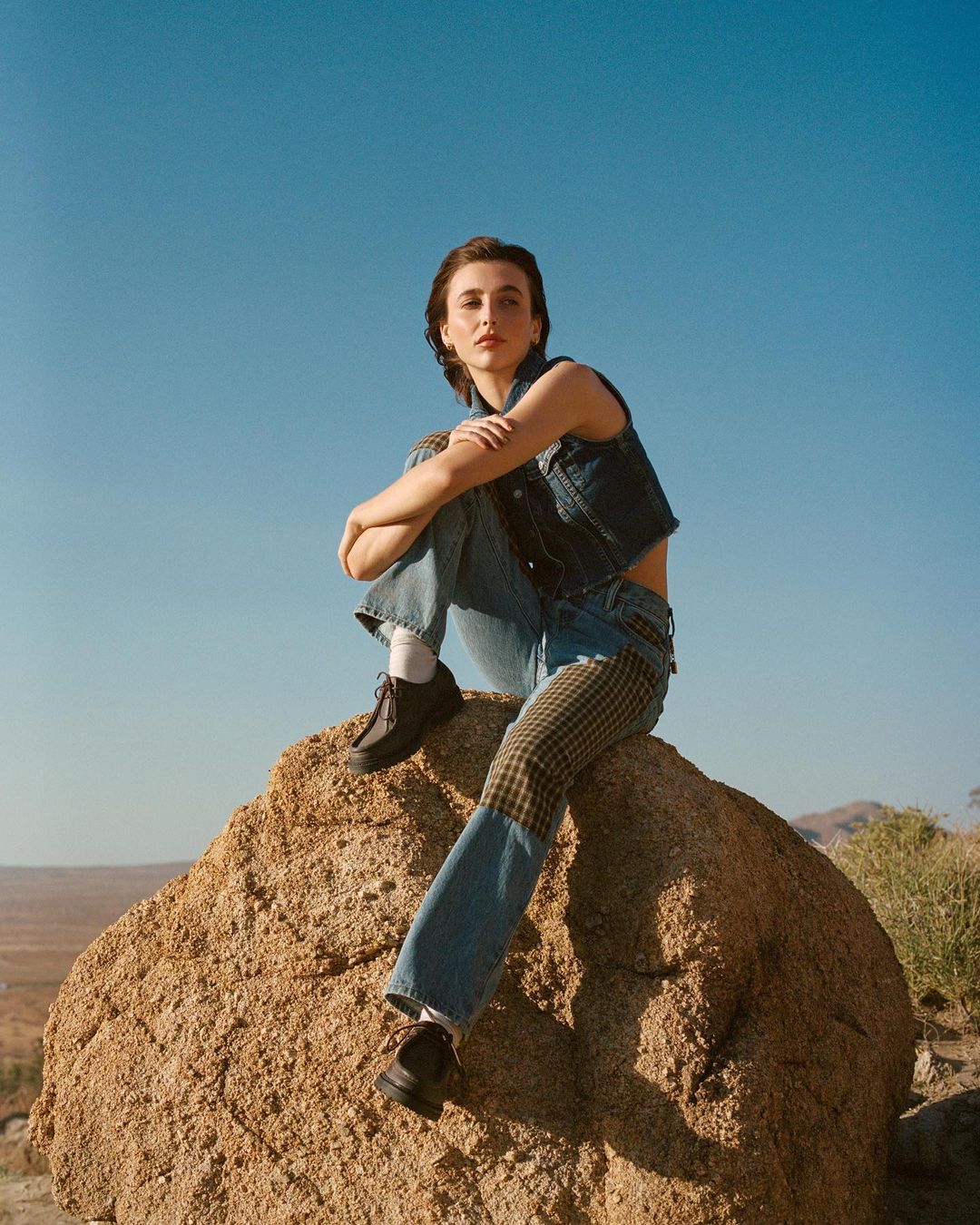 Adult female in denim vest and jeans sits cross-legged on a rock in the desert under a blue sky in a medium outdoor shot.