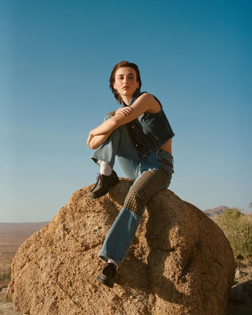 Adult female in denim vest and jeans sits cross-legged on a rock in the desert under a blue sky in a medium outdoor shot.