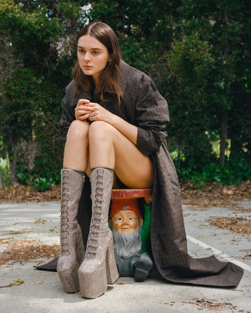 An adult woman sits on a garden gnome figurine wearing platform boots and a trench coat in an outdoor setting with green trees.