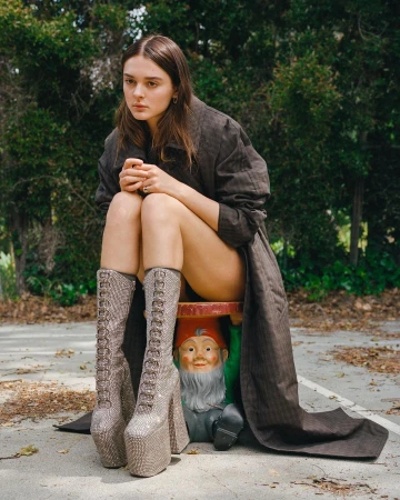 An adult woman sits on a garden gnome figurine wearing platform boots and a trench coat in an outdoor setting with green trees.
