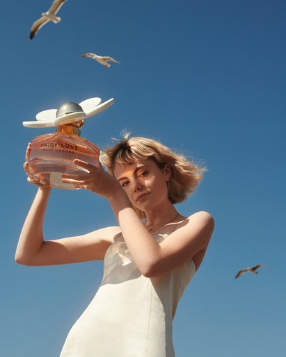 Woman in white dress holding perfume bottle against blue sky with seagulls. Beauty shot for cosmetic advertisement. Summer aesthetic.
