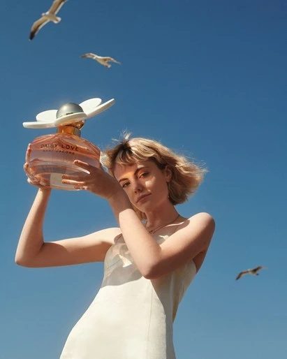 Woman in white dress holding perfume bottle against blue sky with seagulls. Beauty shot for cosmetic advertisement. Summer aesthetic.