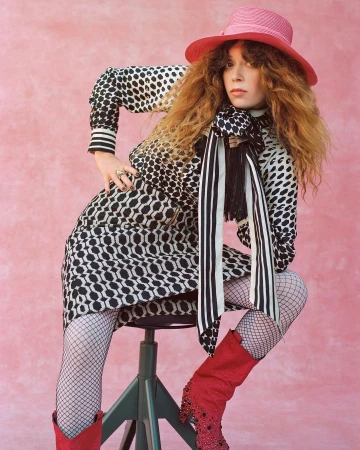 Woman in patterned black and white dress and pink hat sitting on a stool against a pink backdrop
