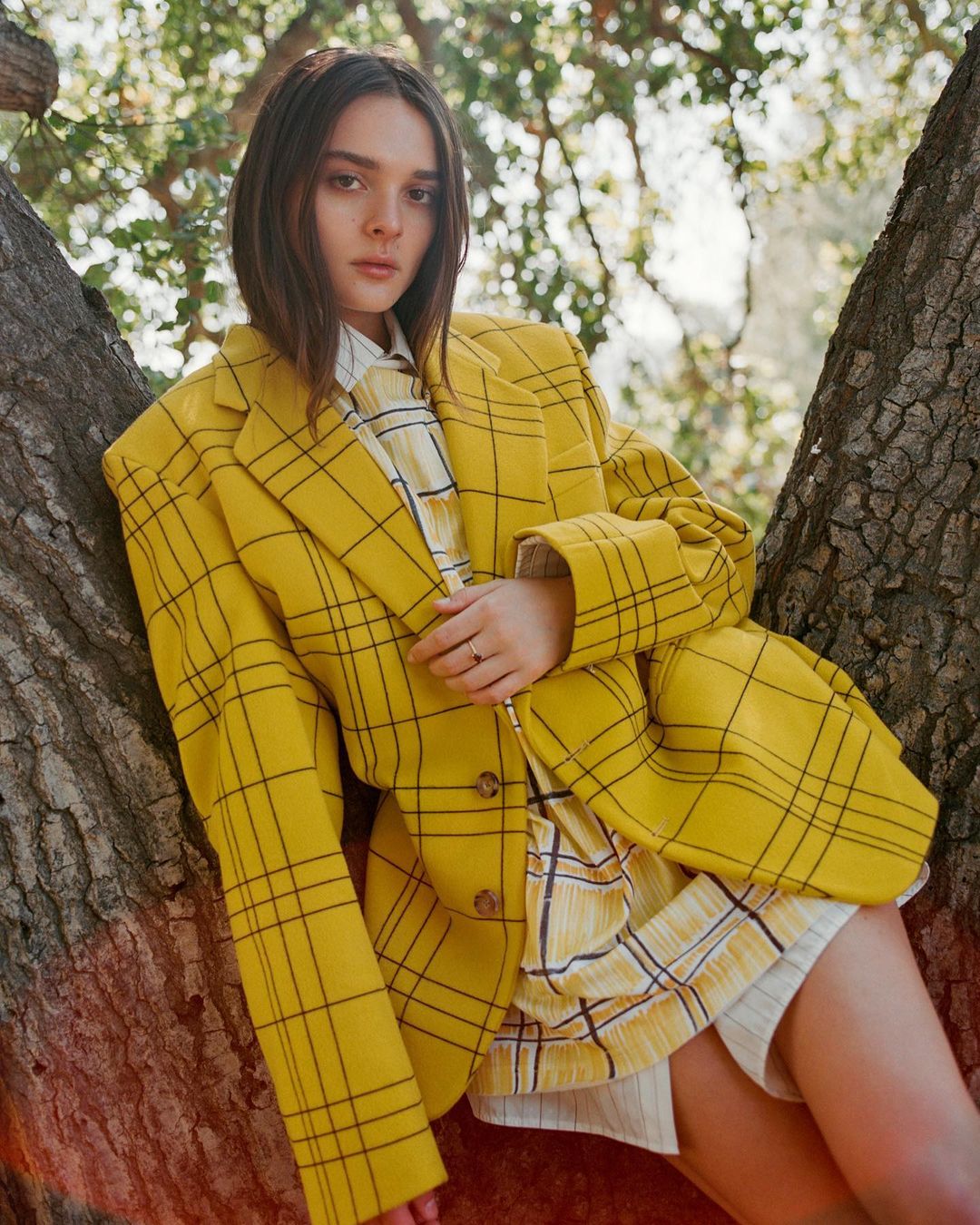 A woman wearing an oversized yellow plaid blazer leans against tree trunks outdoors in natural daylight.