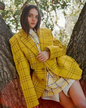 A woman wearing an oversized yellow plaid blazer leans against tree trunks outdoors in natural daylight.