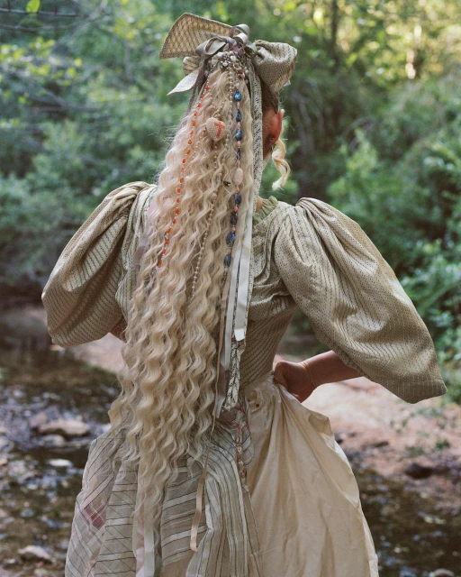 Back view of a woman with long blonde curly hair adorned with ribbons and beads, wearing a vintage dress, standing in a forest stream.