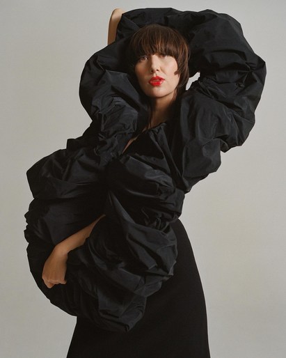 Model in voluminous black puffer dress with arms raised against a plain off-white background