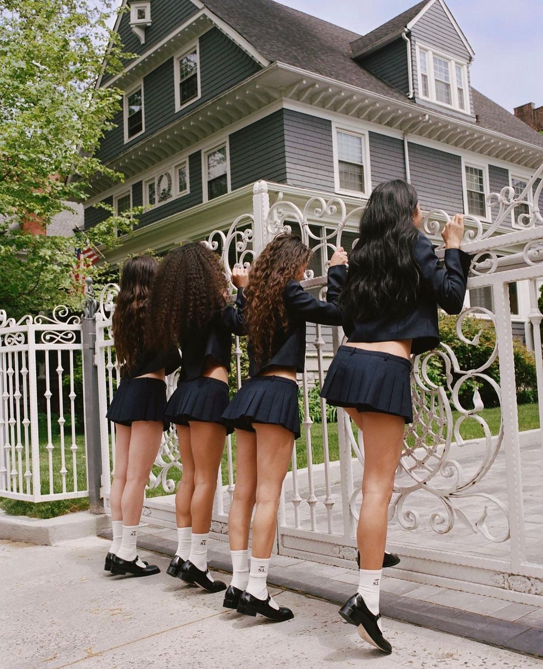 Four young women stand in a row behind a white iron gate in front of a large gray house. The women are wearing matching school uniforms.