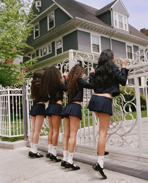 Four young women stand in a row behind a white iron gate in front of a large gray house. The women are wearing matching school uniforms.