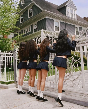 Four young women stand in a row behind a white iron gate in front of a large gray house. The women are wearing matching school uniforms.