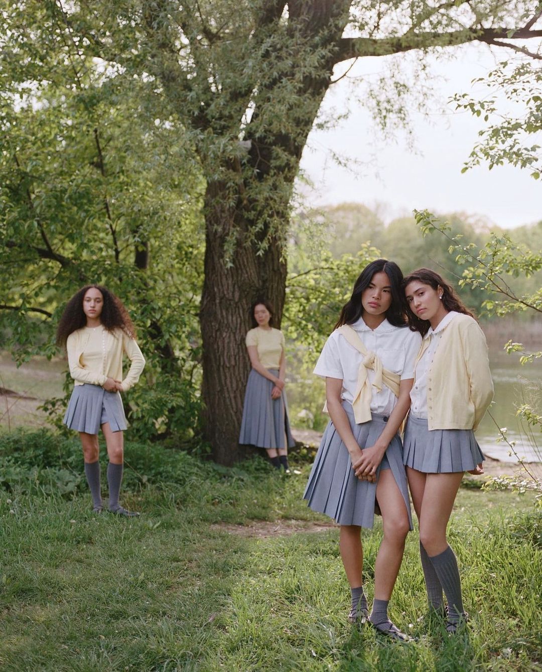 Four young women in pleated skirts and pastel tops pose near a tree in a sunny outdoor setting with lush green grass and foliage.