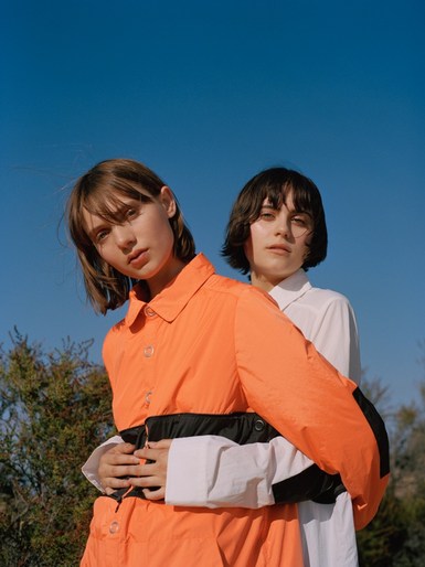Two young women, one in an orange jacket and the other in white, stand closely against a clear blue sky and sparse vegetation.