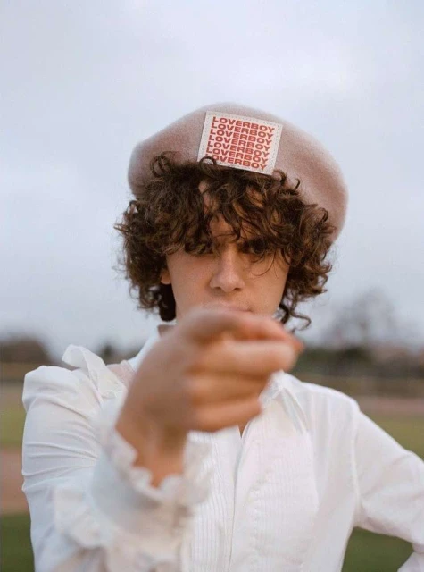 Person in white shirt and beige beret with 'Loverboy' text stands outdoors, pointing towards the viewer with an outstretched hand.