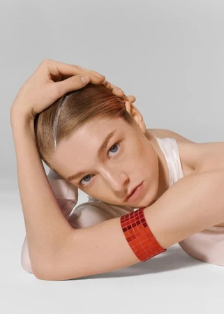 Portrait of a woman with sleek hair and a red mosaic cuff bracelet resting her head on her hands against a grey background.