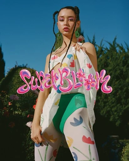 Adult female poses outdoors with decorative tights, floral print top and coat against a backdrop of blue sky and green vegetation.