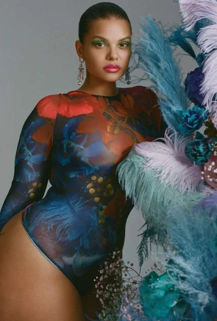 Portrait of an adult female wearing a patterned body suit in a photo studio with feather props and bold green makeup and pink lips.