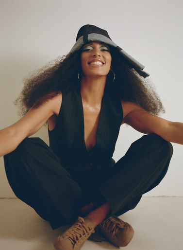 Attractive woman in black vest and pants sitting with crossed legs, smiling with Afro hairstyle and eye makeup, wearing a stylish hat.