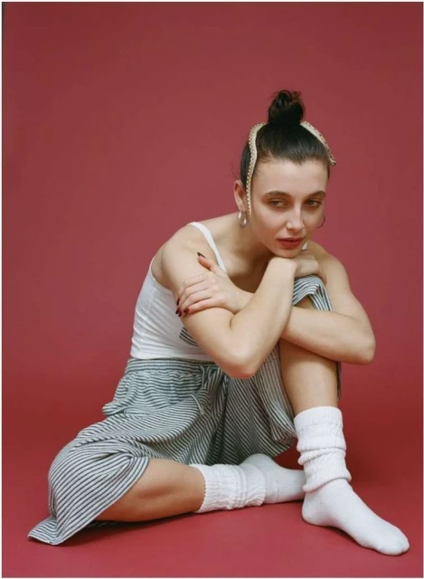 A young woman with a bun hairstyle sits cross-legged on the floor wearing a white tank top, striped pants, white socks, and jewelry.