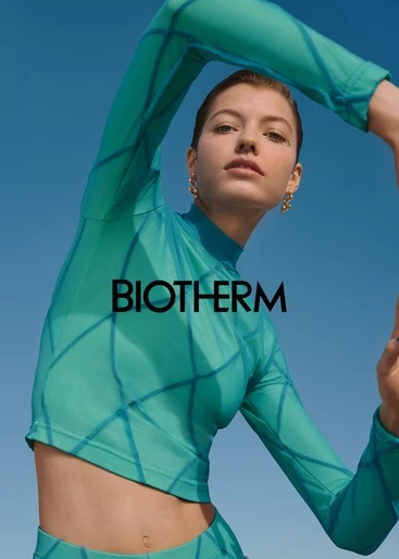 An adult woman wearing teal athletic wear poses against a clear blue sky in an outdoor setting, with the BIOTHERM logo visible.