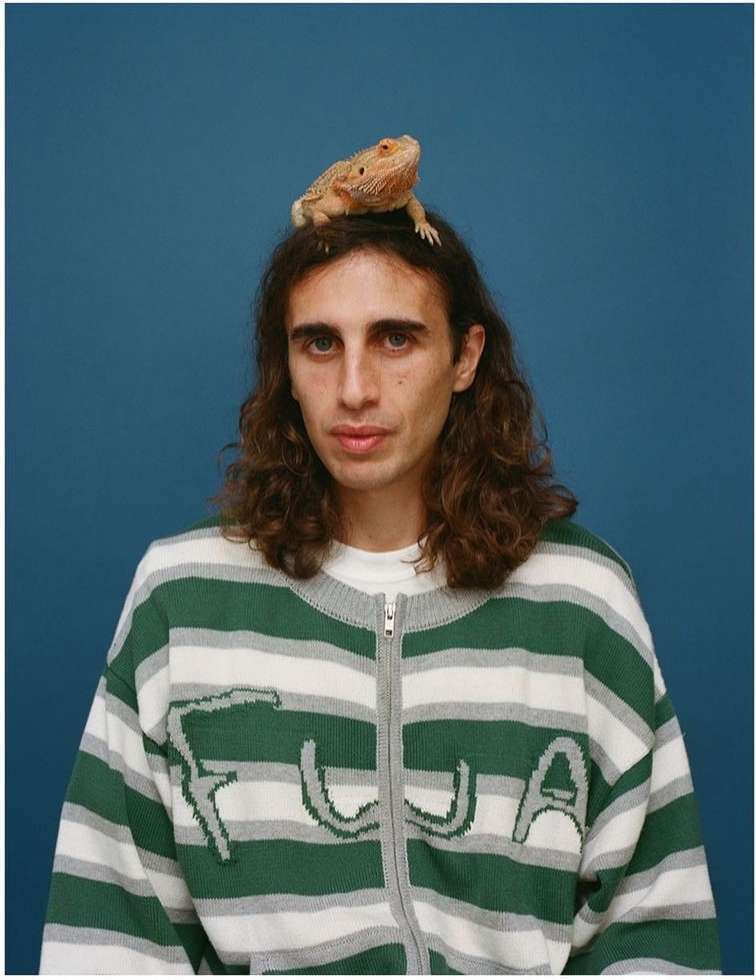 Young man with long brown hair poses with a bearded dragon lizard perched on his head against a solid blue background indoors.