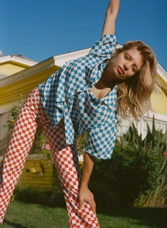 A woman with blonde hair is outdoors in a blue and white checkered shirt and red checkered pants, posing in front of a yellow house.
