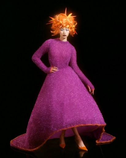 Woman poses in a bright purple, textured long sleeve gown with a high-low hemline. She wears a matching orange spiky hairstyle.