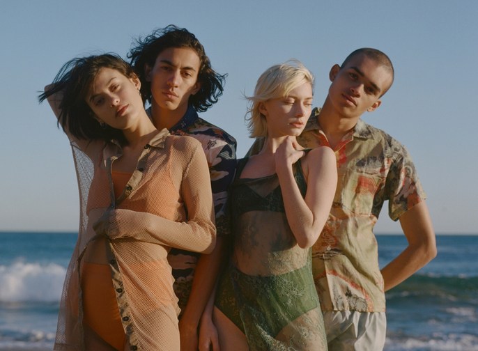 Four young adults pose on a sunny beach with ocean waves; two women wear sheer dresses over swimsuits, and two men wear patterned shirts.
