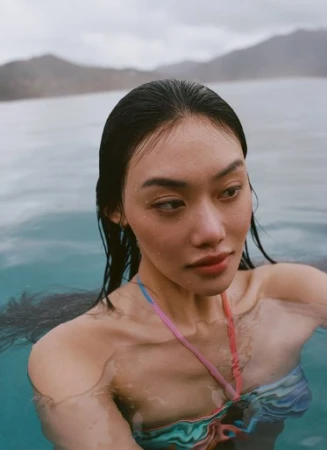 Close-up portrait of an East Asian woman with wet dark hair in turquoise ocean water wearing a colorful printed bikini top.