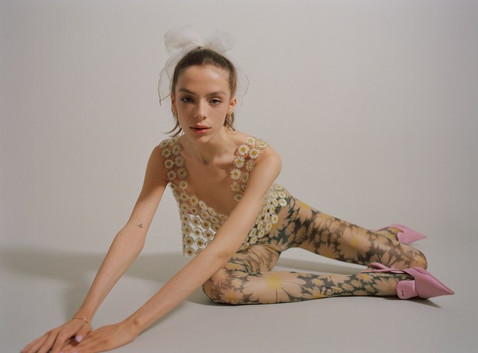A fair-skinned young woman poses while laying down wearing a daisy-patterned top and leggings, complemented by light pink heels and a hair bow.