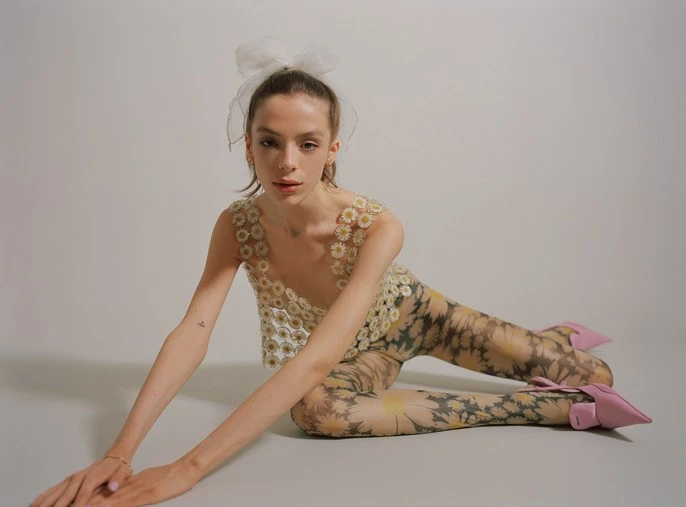 A fair-skinned young woman poses while laying down wearing a daisy-patterned top and leggings, complemented by light pink heels and a hair bow.
