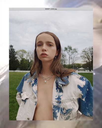 Young adult woman posing outdoors in a tie-dye jacket and necklace, photographed at eye-level in front of a grassy field with trees.