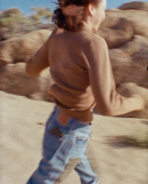 Woman in tan sweater and jeans with patchwork running across a desert landscape with tan rocky formations, captured with motion blur.