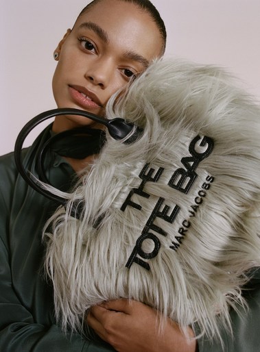 Woman holding a furry, light-grey Marc Jacobs tote bag with black lettering against a pale pink background in a studio setting.