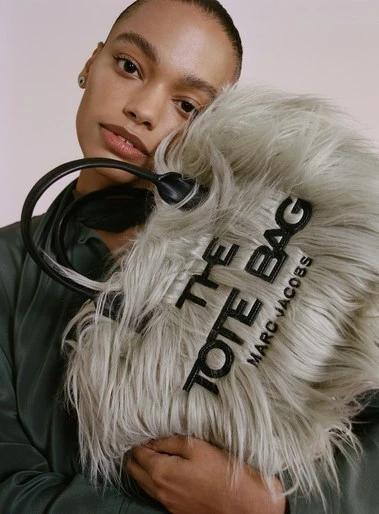 Woman holding a furry, light-grey Marc Jacobs tote bag with black lettering against a pale pink background in a studio setting.