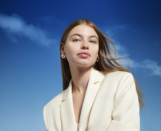 Portrait of an adult woman with long hair wearing a white suit, with silver hoop earrings against a blue sky background with clouds.