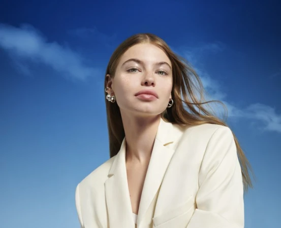 Portrait of an adult woman with long hair wearing a white suit, with silver hoop earrings against a blue sky background with clouds.