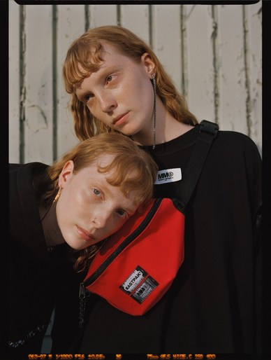 Two fair-skinned young women with reddish-blonde hair pose in a studio. One woman rests her head on the other's shoulder near a red Eastpak bag.