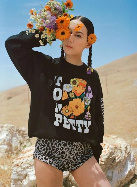 Woman in a Tom Petty sweatshirt poses with flowers on a sunny day. She has braids and is wearing shorts with an animal print.