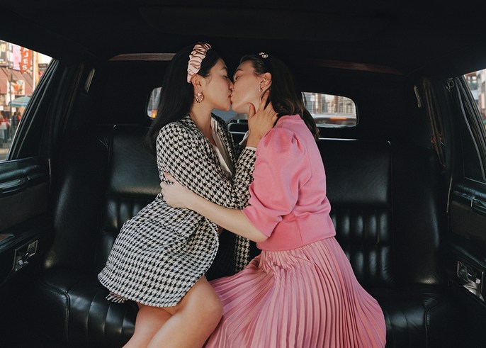 Two women embrace and kiss intimately inside a black car, showcasing a scene of affection with an urban background visible outside.
