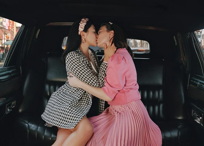 Two women embrace and kiss intimately inside a black car, showcasing a scene of affection with an urban background visible outside.