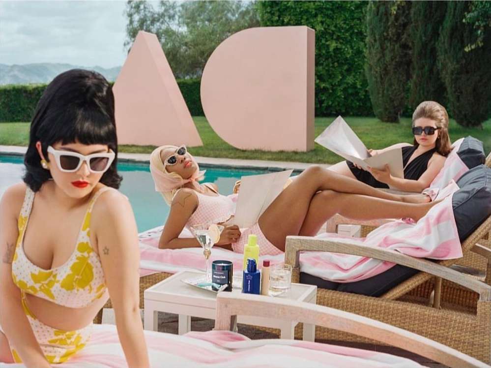 Three women lounge poolside in retro swimwear and sunglasses, beside pink geometric sculptures, embodying a vintage-inspired aesthetic.
