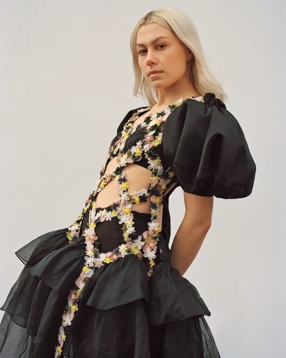 A fair-skinned adult female poses in a semi-profile shot wearing a black dress with floral lattice on a white background in a studio.