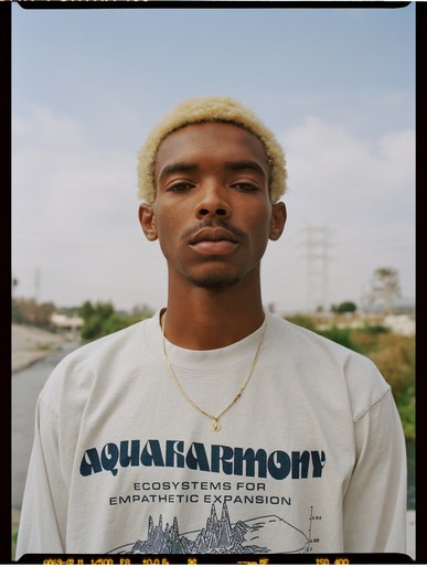 Portrait of a young adult male with dyed blond curly hair, wearing a graphic t-shirt with a necklace, outdoors against a blue sky.