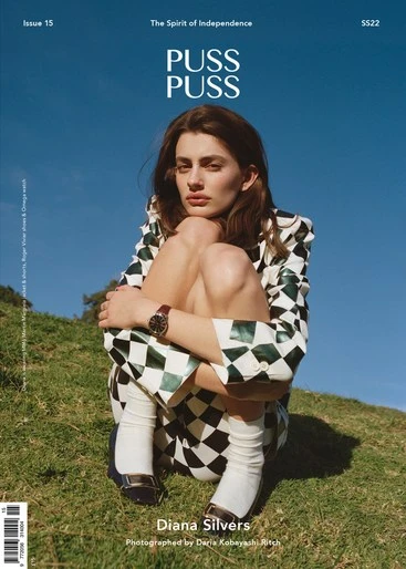 Magazine cover photo of a young woman in a checkered jacket and knee-high socks, posing outdoors against a green hillside and blue sky.