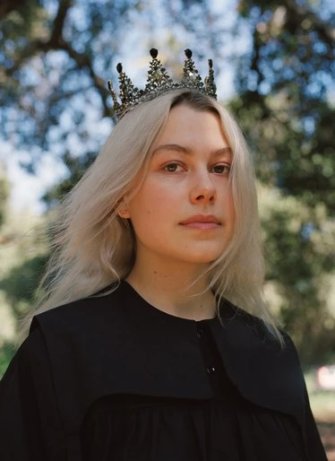 Portrait of a blonde woman with a jeweled crown, wearing a black dress, posing outdoors against a backdrop of green trees and sky.