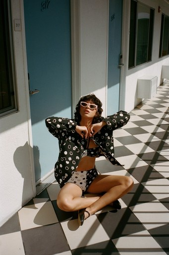 A woman poses in patterned outfit on a tiled floor in front of blue doors, bathed in sunlight, wearing retro sunglasses and open jacket.