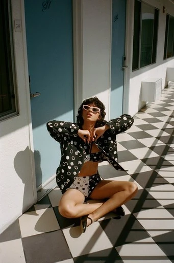 A woman poses in patterned outfit on a tiled floor in front of blue doors, bathed in sunlight, wearing retro sunglasses and open jacket.
